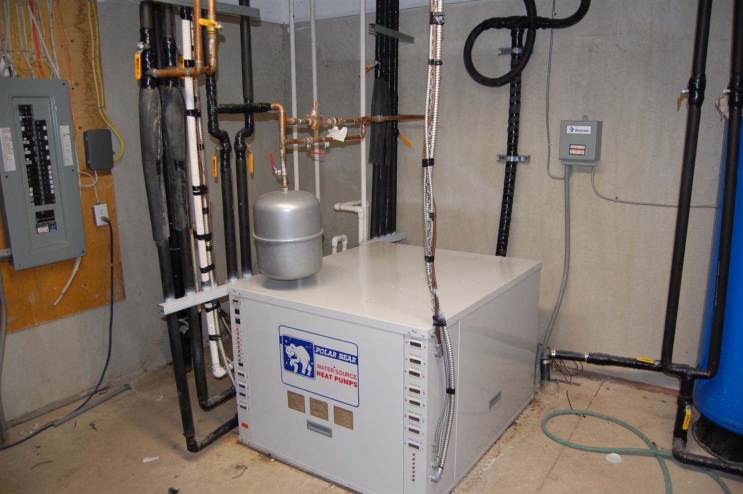 A Polar Bear water source heat pump installed in a mechanical room, connected to insulated loop piping, an expansion tank and an electrical panel.